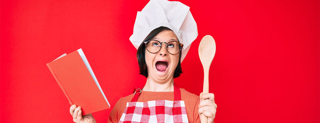 Brunette woman with down syndrome wearing professional baker apron reading cooking recipe book