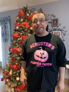 Meet Jorge. Wearing a colorful black and pink tshirt and glasses, Jorge is a brunette. He is standing in front of a Christmas tree decorated primarily in red. 