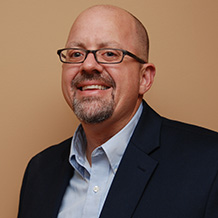 Mark smiling, wearing glasses. Dressed in a light blue shirt with dark suit jacket. Partially balding.