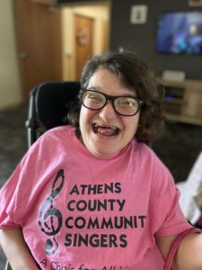 Deidra is wearing black rimmed glasses and has dark hair. She is smiling and wearing a pink t-shirt. 