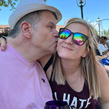 Frannie and Lindsey. Lindsey is in a black and white tank top. She has blonde hair. Frannie is wearing a hat and a pink t-shirt. He is giving Lindsey a kiss on the cheek.