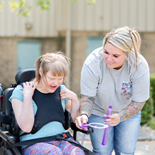 DSP helping an individual with disabilities to have fun blowing bubbles out side. Both are smiling. DSP has blond hair and is wearing a gray t-shirt. The individual is laughing wearing a teal t-shirt