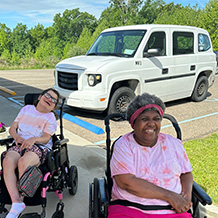 Two people who live at Echoing Hills are shown sitting in their wheelchairs with the adaptable van in the background that takes them into the community. The van is white with the Corporate blue logo. One person is wearing a pink flowered shirt with pink pants. Both are smiling.