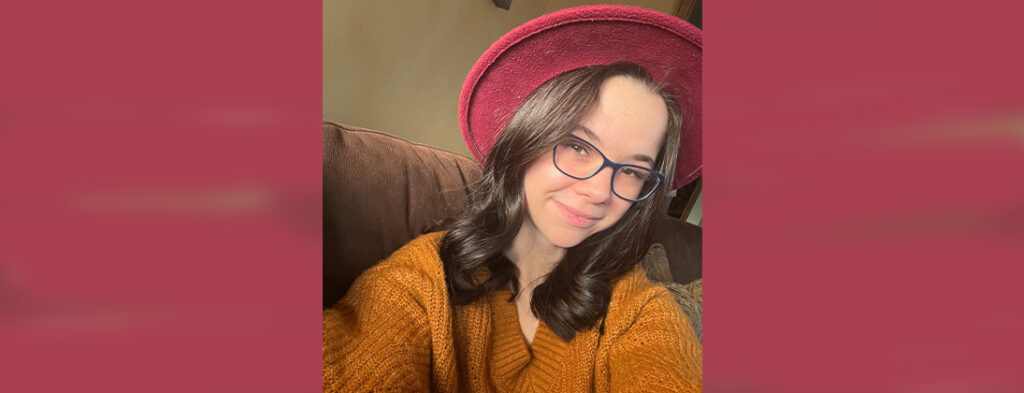 Meet Harley Comisford. Beautiful young lady smiling with her head tilted. She wears glasses and has medium length brown hair. She is wearing a Rose colored hat with a large brim. She is wearing a gold sweater and is sitting in a brown chair.